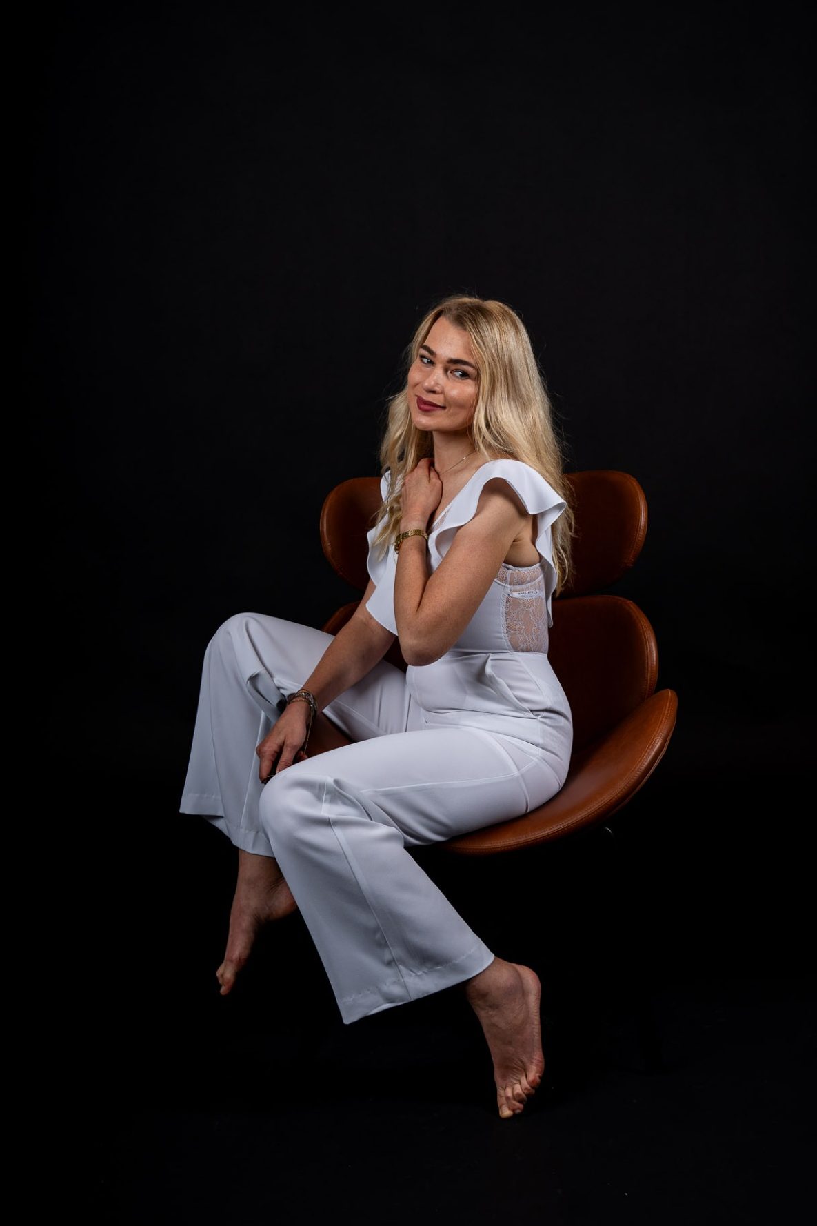 Frau im weißen Jumpsuit sitzt auf Stuhl vor schwarzem Hintergrund